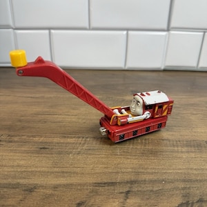 May include: A red toy crane vehicle featuring a character face with white and grey details. The crane has a long, articulated red arm with a yellow tip, a white roof, and yellow diagonal stripes on the side of the base. The toy is made of metal.