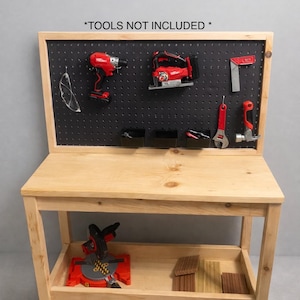 Peut inclure: Un établi en bois avec un panneau perforé noir à l'arrière et une étagère inférieure. Le panneau perforé affiche divers outils électriques rouges et des lunettes de sécurité. Le texte "*OUTILS NON INCLUS*" est en haut.