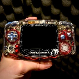 Puede incluir: Una consola portátil Game Boy Advance transparente con temática de Spider-Man. La consola presenta una pantalla negra con un diseño de telaraña blanca y el texto "Spider-Man" y "Game Boy Advance". Los botones son rojos y azules.