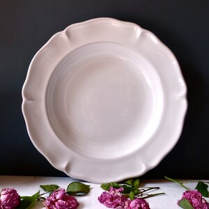 May include: A large, off-white ceramic plate with a scalloped edge. The plate is centered against a dark background, with dried pink roses and green leaves scattered around the base. The plate's design is simple and elegant.