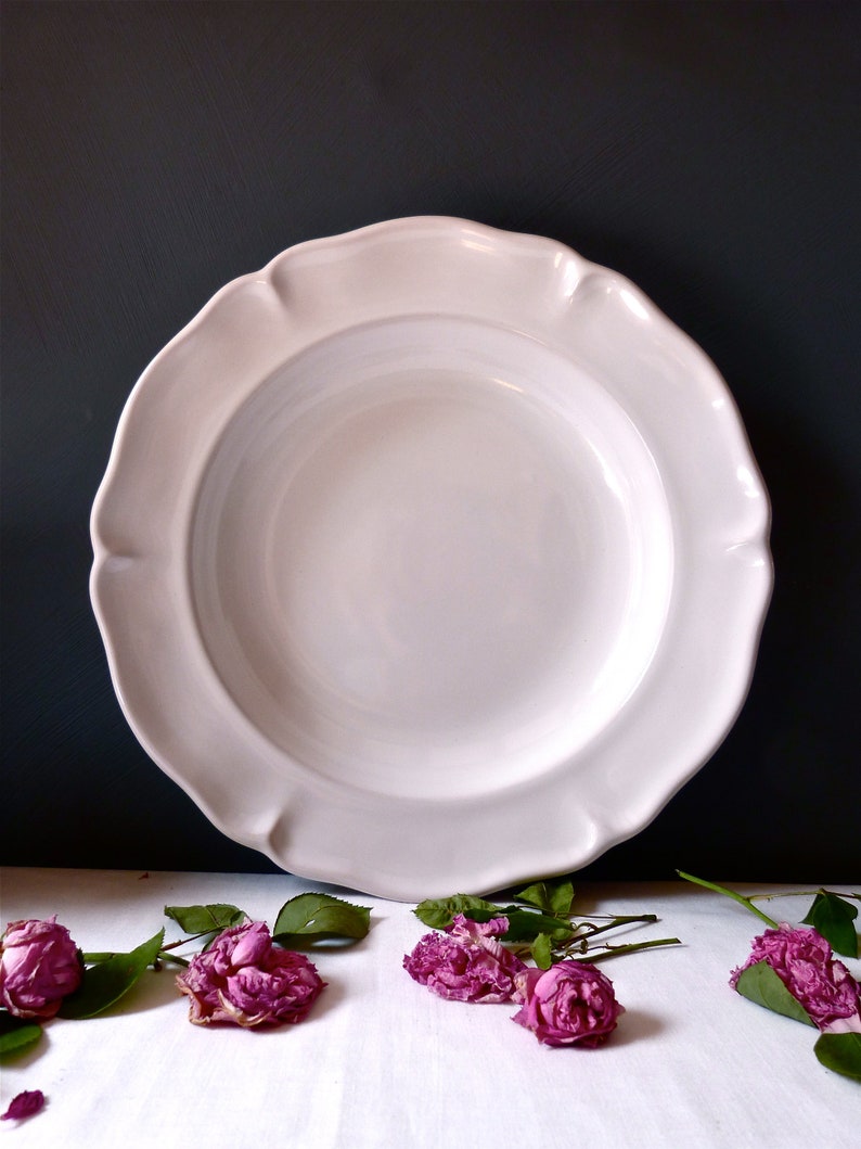 May include: A large, off-white ceramic plate with a scalloped edge. The plate is centered against a dark background, with dried pink roses and green leaves scattered around the base. The plate's design is simple and elegant.