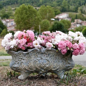 May include: A rectangular, footed ceramic planter with a weathered, blue-grey finish and an intricate, raised lattice and floral relief pattern. The planter is overflowing with a dense arrangement of light pink, dark pink, and white roses.
