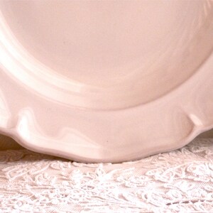 May include: A close-up of a pale pink ceramic plate with a scalloped edge. The plate has a smooth, glossy finish and sits on a white lace tablecloth. The plate's design is simple and elegant.