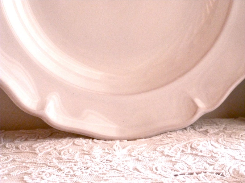 May include: A close-up of a pale pink ceramic plate with a scalloped edge. The plate has a smooth, glossy finish and sits on a white lace tablecloth. The plate's design is simple and elegant.