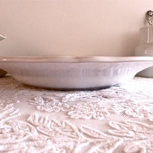 May include: A white ceramic serving bowl with a slightly raised rim and a light beige edge. A white pitcher and a gravy boat are in the background. The bowl sits on a white lace tablecloth.