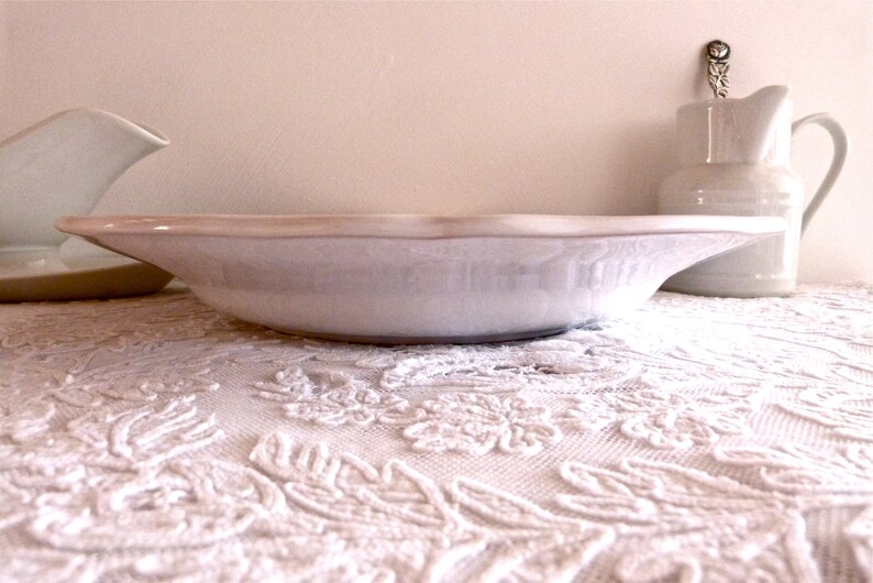 May include: A white ceramic serving bowl with a slightly raised rim and a light beige edge. A white pitcher and a gravy boat are in the background. The bowl sits on a white lace tablecloth.