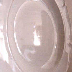 May include: A white ceramic plate with a scalloped edge. The plate has a wide rim and a slightly recessed center. The surface is smooth and glossy, reflecting light. The plate is likely for serving food.