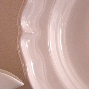 May include: Close-up of a white ceramic plate with a scalloped edge. The plate has a glossy finish and a curved design. The image shows a portion of the plate, highlighting its elegant shape and texture. The plate is likely part of a dinnerware set.