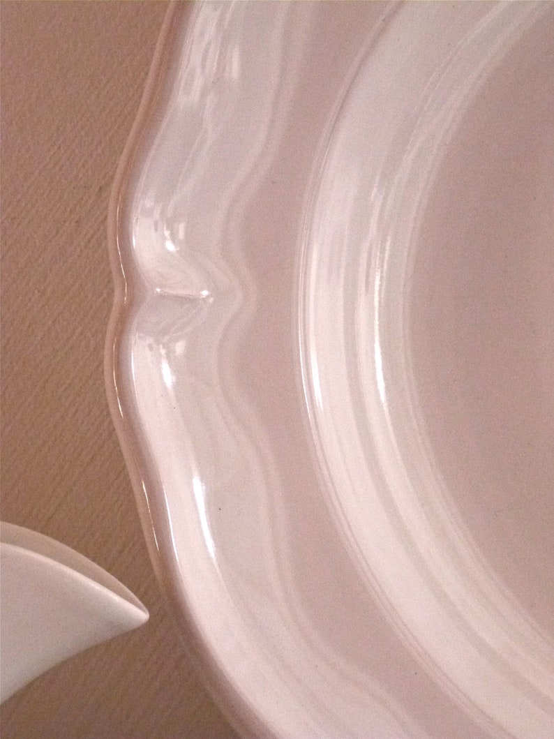 May include: Close-up of a white ceramic plate with a scalloped edge. The plate has a glossy finish and a curved design. The image shows a portion of the plate, highlighting its elegant shape and texture. The plate is likely part of a dinnerware set.