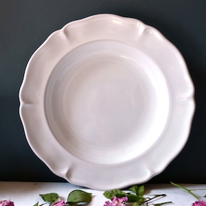 May include: A large, white ceramic plate with a scalloped rim. The plate has a wide, shallow bowl and a smooth, glossy finish. The plate is a classic design, suitable for serving a variety of dishes. The background is a dark teal color.