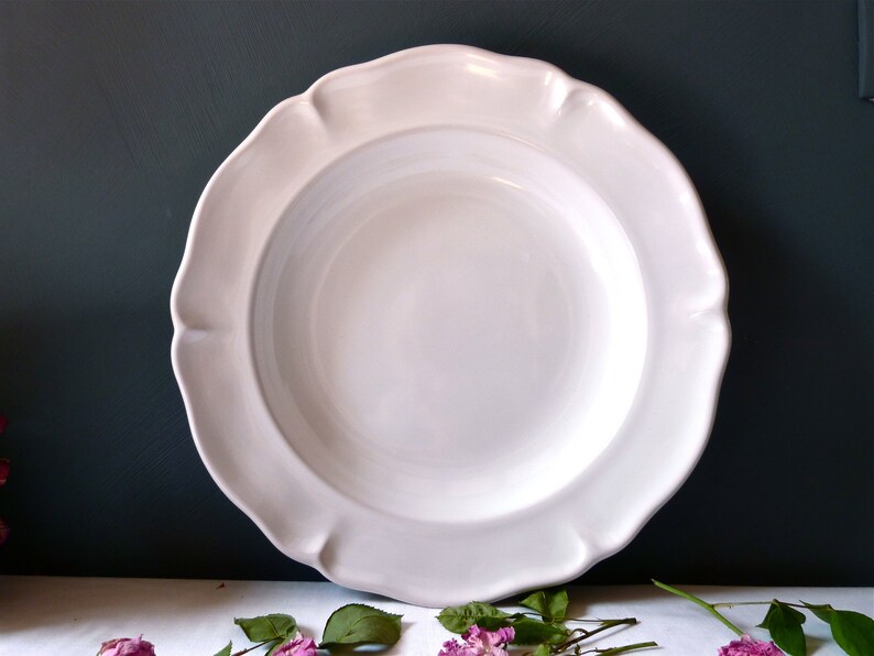 May include: A large, white ceramic plate with a scalloped rim. The plate has a wide, shallow bowl and a smooth, glossy finish. The plate is a classic design, suitable for serving a variety of dishes. The background is a dark teal color.