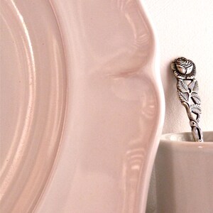 May include: A close-up of a pale pink plate with a scalloped edge. A decorative silver spoon with a rose design rests on the edge of a white cup. The spoon features intricate floral details.