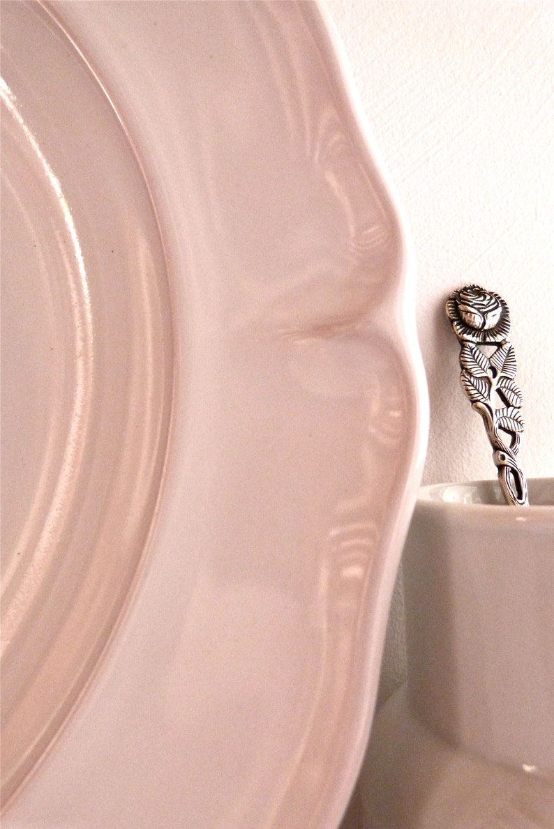 May include: A close-up of a pale pink plate with a scalloped edge. A decorative silver spoon with a rose design rests on the edge of a white cup. The spoon features intricate floral details.