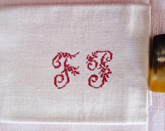 May include: White linen towel with red embroidered initials "F B". The initials are in a decorative, floral style. The towel is folded and resting on a pink and white checkered surface. A horn-like object is visible on the right side.