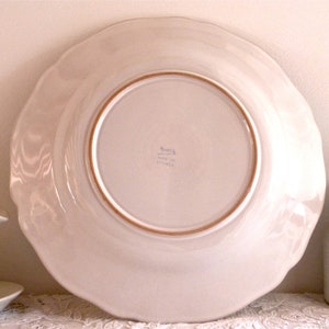May include: A large, off-white ceramic plate with a scalloped edge. The underside of the plate is visible, showing a light brown rim and the words "Made in France". A decorative spoon and small pitcher are also in the image.