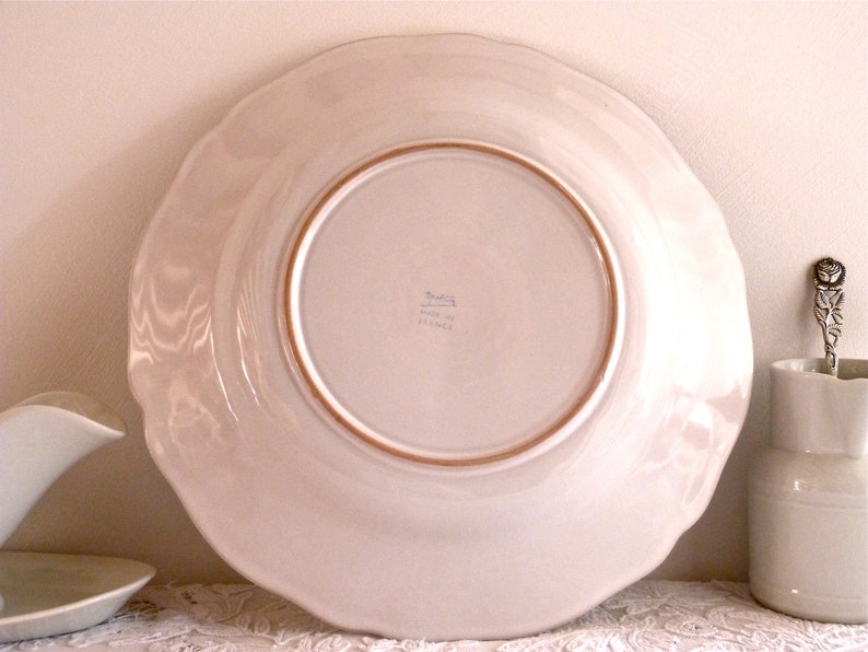May include: A large, off-white ceramic plate with a scalloped edge. The underside of the plate is visible, showing a light brown rim and the words "Made in France". A decorative spoon and small pitcher are also in the image.