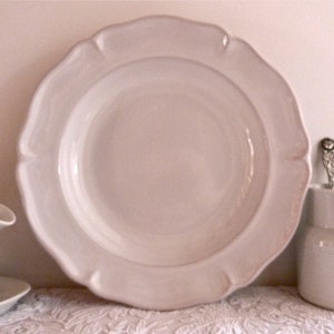 May include: A collection of white ceramic tableware, including a large plate with a scalloped edge, a small pitcher, and a gravy boat. The plate is the focal point, with the other pieces arranged around it. The items are on a white lace tablecloth.