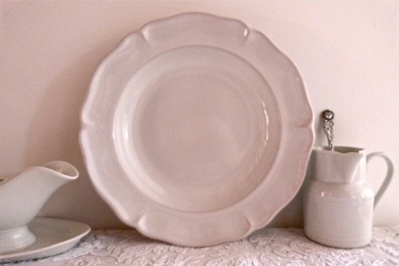 May include: A collection of white ceramic tableware, including a large plate with a scalloped edge, a small pitcher, and a gravy boat. The plate is the focal point, with the other pieces arranged around it. The items are on a white lace tablecloth.