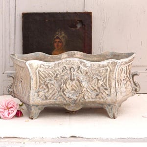 May include: An aged, rectangular planter with detailed relief patterns. The planter has a distressed white and blue paint finish, with decorative handles and four small feet. A pink rose and a vintage painting are in the background.