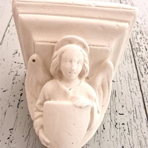 May include: Off-white angel wall shelf. The angel has a halo and wings, holding a shield. The shelf has a rectangular top and is made of a solid material. The background is a distressed white and light blue wood.