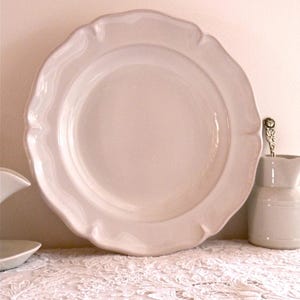 May include: A large, off-white ceramic plate with a scalloped edge. The plate is round with a slightly raised center. A small, matching pitcher and a spoon are visible to the right. The plate is sitting on a white lace tablecloth.