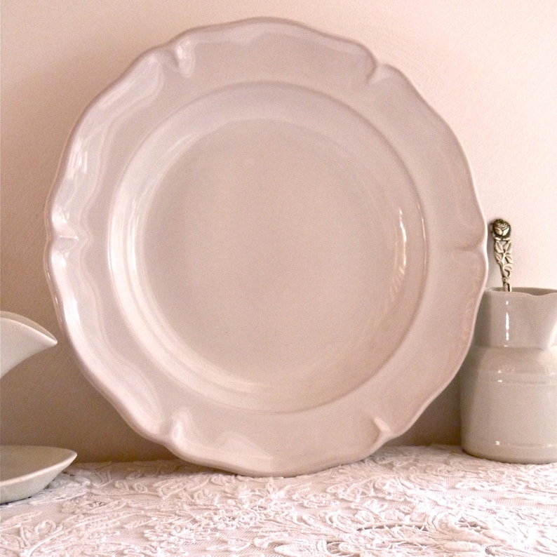 May include: A large, off-white ceramic plate with a scalloped edge. The plate is round with a slightly raised center. A small, matching pitcher and a spoon are visible to the right. The plate is sitting on a white lace tablecloth.
