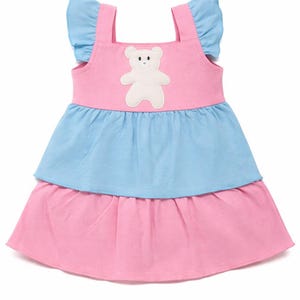 Pode incluir: Um vestido de bebê em camadas rosa e azul com uma aplicação de urso de pelúcia branco. O vestido tem mangas com babados e alças. A camada inferior é rosa, a camada do meio é azul e a parte superior é rosa.