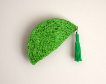 May include: A bright green, semi-circular crochet clutch with a matching tassel zipper pull. The handmade bag is a vibrant accessory, perfect for carrying essentials. The bag is made of a textured yarn.