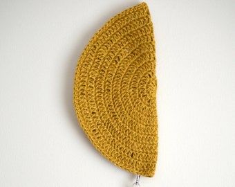 May include: Mustard yellow crocheted wall hanging in a half-circle shape. A matching tassel hangs from the bottom, attached with a silver-colored clasp. The wall hanging is displayed against a white background.