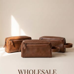Peut inclure: Trois trousses de toilette en cuir marron avec fermetures éclair et une poignée sur l'une d'elles. Les sacs sont disposés sur une surface claire. Le mot "WHOLESALE" est imprimé en marron foncé au bas de l'image.