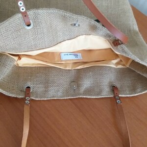 May include: A beige burlap tote bag with brown leather handles and accents. The bag has a light yellow interior lining and a magnetic snap closure. The handles are attached with silver-colored grommets.