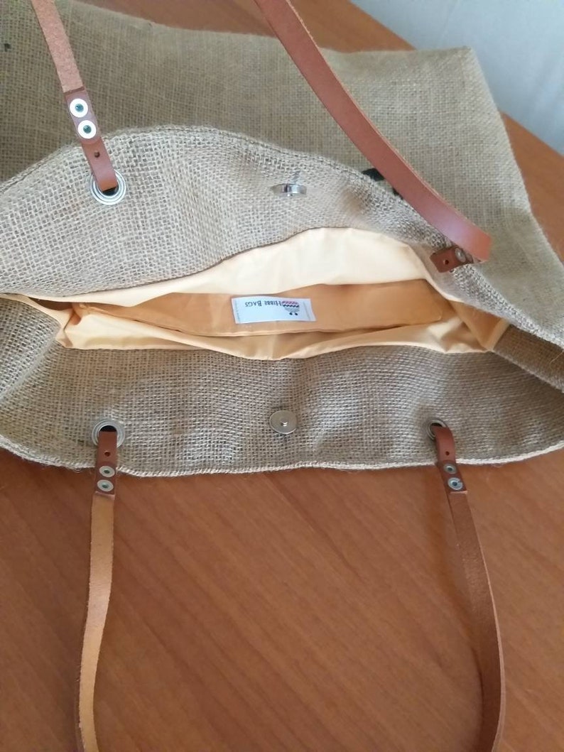 May include: A beige burlap tote bag with brown leather handles and accents. The bag has a light yellow interior lining and a magnetic snap closure. The handles are attached with silver-colored grommets.