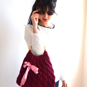 May include: A burgundy cable-knit handbag with bamboo handles and a pink satin bow. The bag has a rounded shape and a textured pattern. The woman is wearing a white sweater and sunglasses.