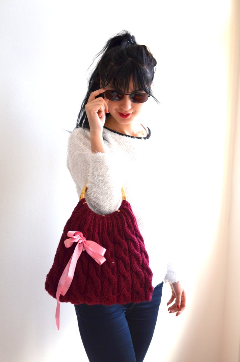 May include: A burgundy cable-knit handbag with bamboo handles and a pink satin bow. The bag has a rounded shape and a textured pattern. The woman is wearing a white sweater and sunglasses.
