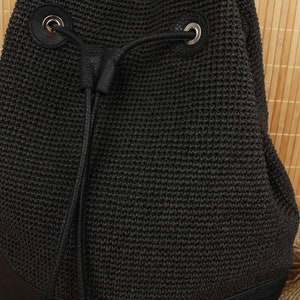 May include: Black woven drawstring backpack with leather accents. The bag features a textured, knitted design and a drawstring closure with silver-toned metal eyelets. The bag's base is made of black leather.