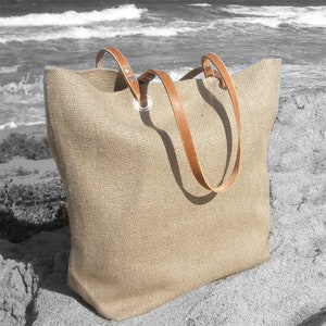 May include: A large, beige burlap tote bag with brown leather handles. The bag has a rectangular shape and is sitting on a sandy surface near the ocean. The handles are attached with silver metal grommets.