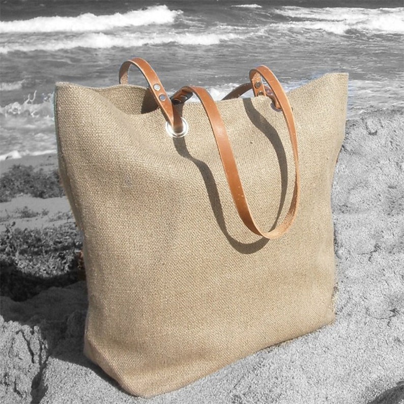 May include: A large, beige burlap tote bag with brown leather handles. The bag has a rectangular shape and is sitting on a sandy surface near the ocean. The handles are attached with silver metal grommets.