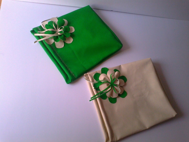 May include: Two square gift boxes, one green and one beige, each adorned with a decorative flower. The flowers feature green and cream-colored petals and ribbon accents, adding a touch of elegance to the wrapped presents.