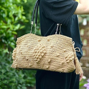 May include: A beige crocheted handbag with a textured pattern and black leather handles. The bag features a decorative tassel and chain straps. The bag is held by a person in a black shirt.