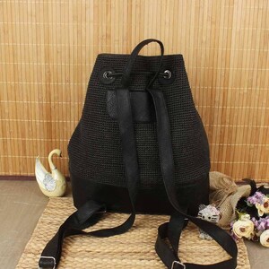 May include: Black backpack with a woven texture and leather accents. The backpack has adjustable straps and a drawstring closure. The bottom of the bag is made of solid black leather. The bag is sitting on a woven mat.