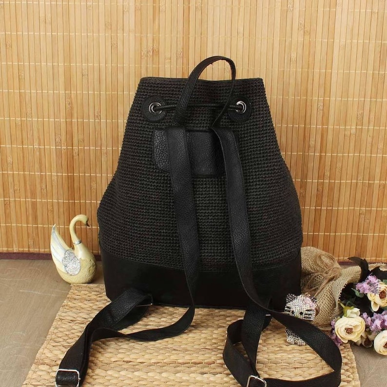 May include: Black backpack with a woven texture and leather accents. The backpack has adjustable straps and a drawstring closure. The bottom of the bag is made of solid black leather. The bag is sitting on a woven mat.