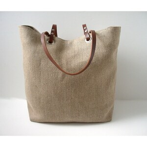 May include: A beige tote bag with brown leather handles. The bag is made of a woven fabric and has a rectangular shape. The handles are attached to the bag with silver-colored metal grommets.