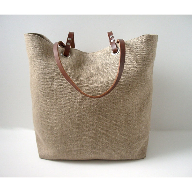 May include: A beige tote bag with brown leather handles. The bag is made of a woven fabric and has a rectangular shape. The handles are attached to the bag with silver-colored metal grommets.