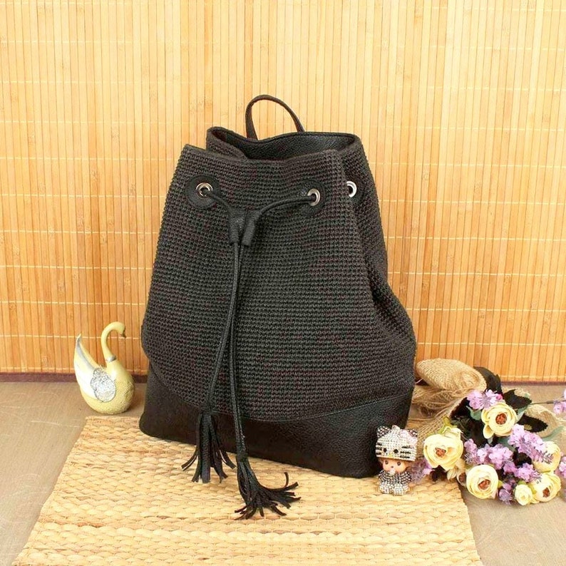 May include: Black backpack with a drawstring closure and leather accents. The bag features a woven texture on the upper portion and a solid leather base. A small handle is at the top, and the drawstring has tassels.