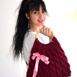 May include: A burgundy knitted handbag with a cable knit design and a bamboo handle. The bag features a pink satin ribbon bow. The bag is a stylish accessory.