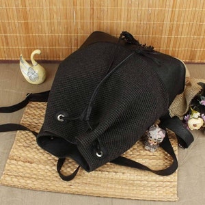 May include: Black backpack with a woven body and leather accents. The drawstring closure features metal grommets. The bag has adjustable straps and a small tassel. A small cat figurine and a swan figurine are visible.