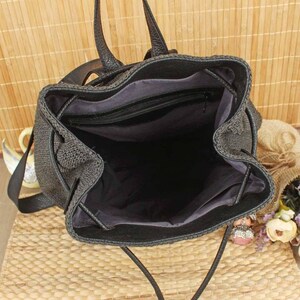 May include: Black woven backpack with a drawstring closure and leather straps. The interior is black with a zippered pocket. The bag is open, revealing the inside. The bag is sitting on a woven surface.