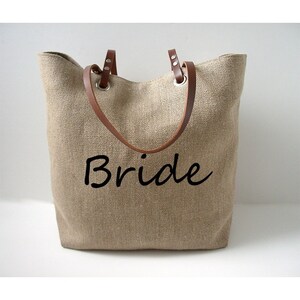 May include: A beige burlap tote bag with brown leather handles and the word "Bride" in black script. The bag has a simple, open design, perfect for carrying items.