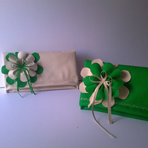 May include: Two fabric clutches, one cream and one green, each adorned with a large flower. The flowers feature green and cream petals, with ribbon accents. The clutches are rectangular and appear to be made of a textured fabric.