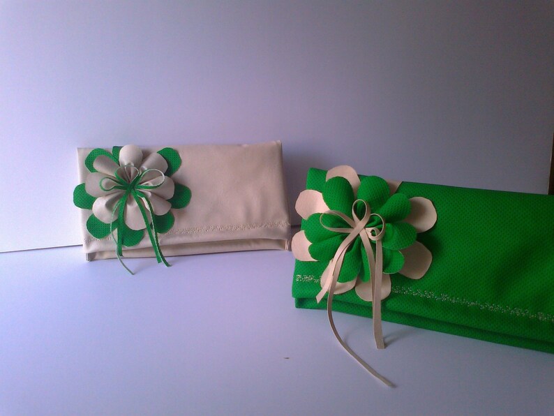 May include: Two fabric clutches, one cream and one green, each adorned with a large flower. The flowers feature green and cream petals, with ribbon accents. The clutches are rectangular and appear to be made of a textured fabric.